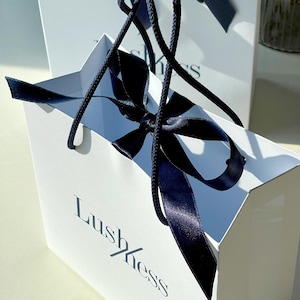 May include: Two white gift bags with navy blue ribbon handles and bows. The bags have the word "Lushness" printed on the front in a stylish font. The bags are sitting on a light-colored surface.