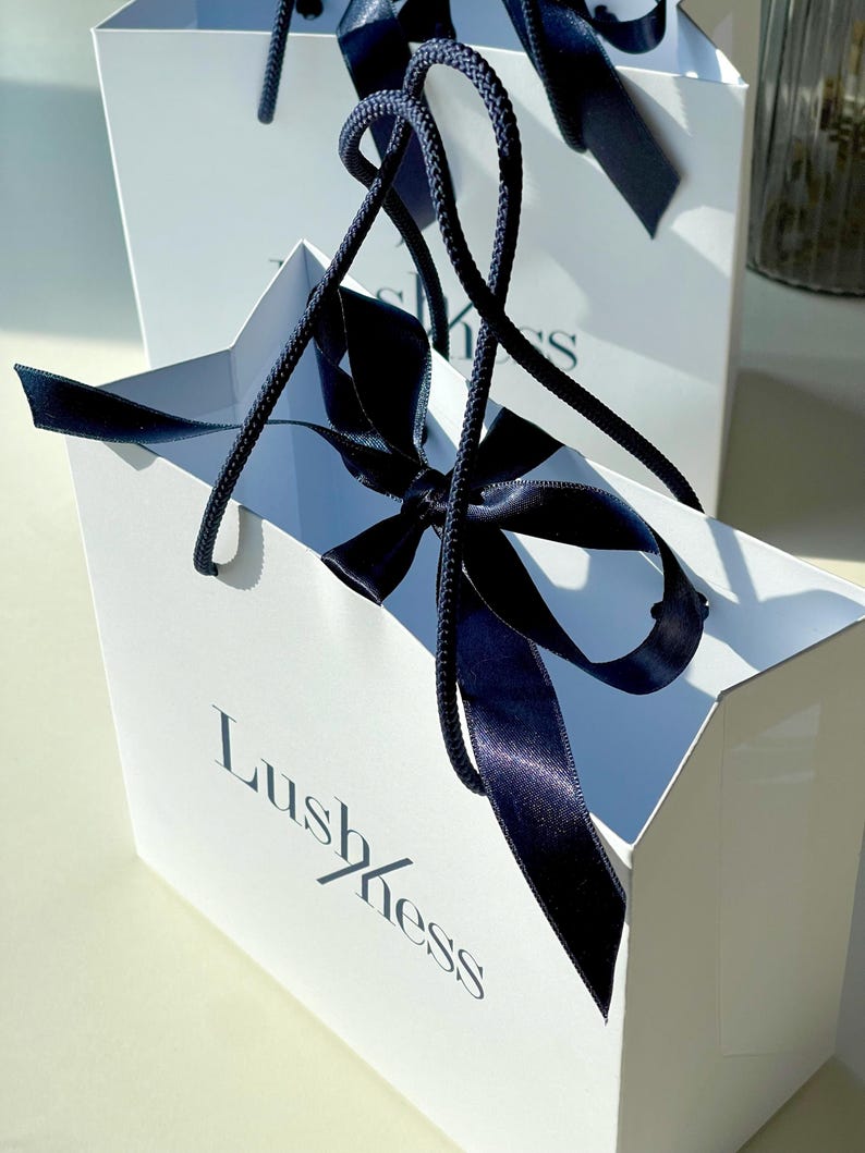 May include: Two white gift bags with navy blue ribbon handles and bows. The bags have the word "Lushness" printed on the front in a stylish font. The bags are sitting on a light-colored surface.
