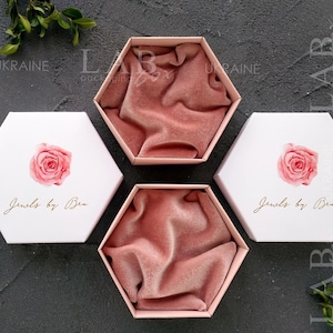 May include: Three hexagonal boxes with pink velvet interiors and white lids. The lids feature a watercolor rose design and the text "Jewels by Ben".