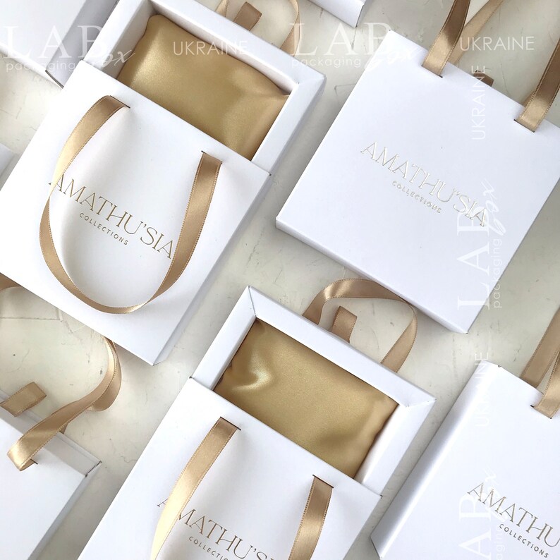 K&ouml;nnte beinhalten: Wei&szlig;e Geschenkboxen mit goldenen Schleifenb&auml;ndern und einem goldenen Satin-Innenfutter. Die Boxen tragen den Schriftzug "Amathusia Collections" auf der Vorderseite.
