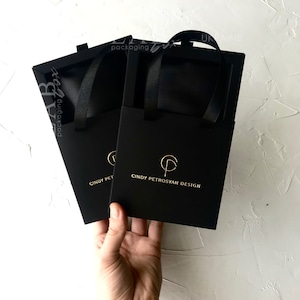 Puede incluir: Dos bolsas de regalo negras con letras doradas y asas de cinta. Las bolsas tienen el texto "CINDY PETROSYAN DESIGN" y un logotipo. Las bolsas son sostenidas por una mano sobre un fondo blanco.