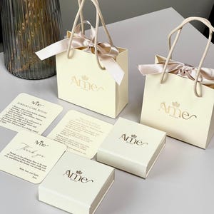 Può includere: Sacchetti e scatole regalo color crema con il logo "Ame" e un motivo floreale. Le borse hanno fiocchi di nastro e manici in corda. Le carte di accompagnamento descrivono la cura dei gioielli e un rituale di benvenuto. Gli articoli sono su una superficie chiara.