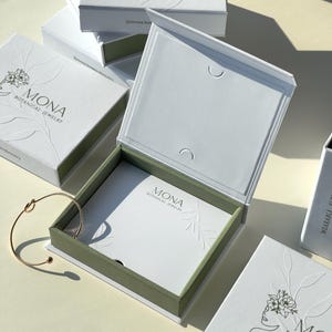 May include: White jewelry boxes with green trim and embossed floral designs. The boxes are labelled "MONA BOTANICAL JEWELRY." A gold-coloured bracelet rests near an open box. The boxes are stacked on a light-coloured surface.