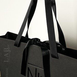 Puede incluir: Bolsa de compras negra con asas de cinta negra y un lazo negro. La bolsa tiene el texto "LAB packaging" y "UKRAINE" impreso en el lateral. La bolsa tambi&eacute;n tiene un logotipo negro con la letra "N" impresa en la parte delantera.