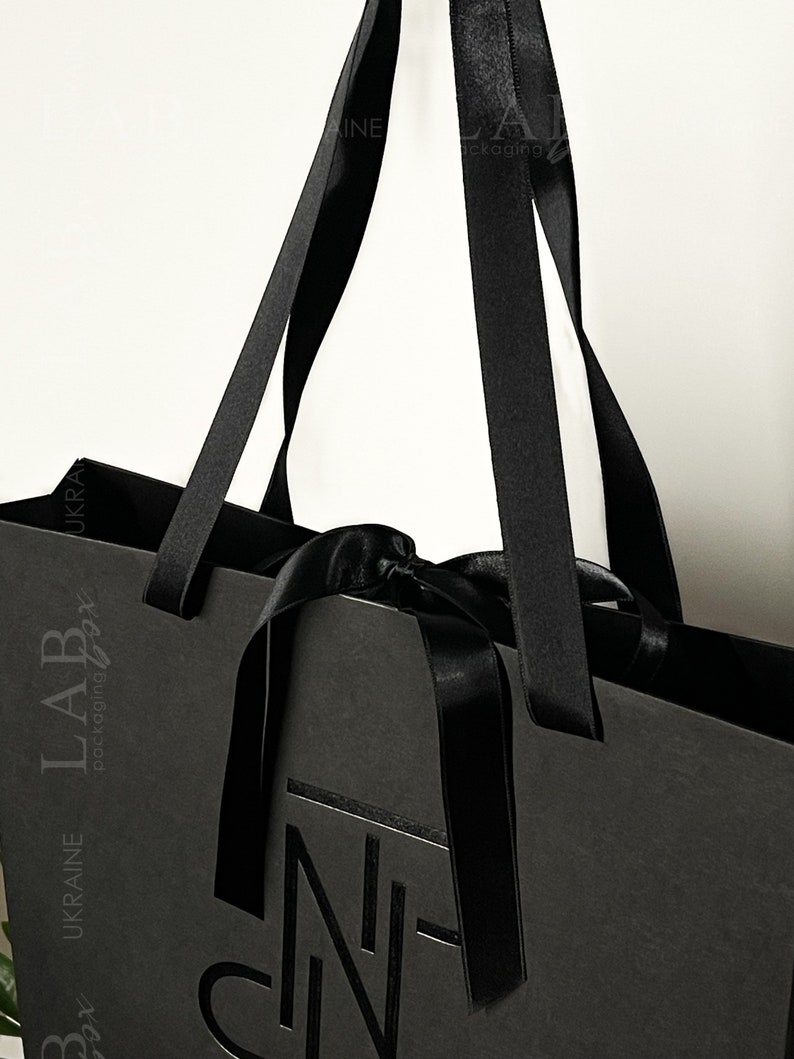 Puede incluir: Bolsa de compras negra con asas de cinta negra y un lazo negro. La bolsa tiene el texto "LAB packaging" y "UKRAINE" impreso en el lateral. La bolsa tambi&eacute;n tiene un logotipo negro con la letra "N" impresa en la parte delantera.