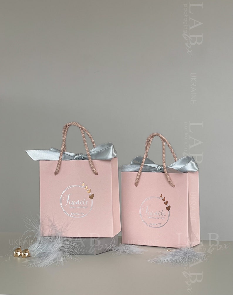 May include: Two pink gift bags with silver handles and bows. The bags have a silver logo with the text "Fiocci" and a circle of gold hearts. The bags are sitting on a white surface with white feathers.