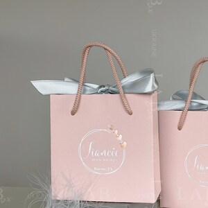 May include: Two pink gift bags with white rope handles and silver satin bows. The bag on the left has a logo that says "Fiancee Bridal Boutique, Boerne, TX".