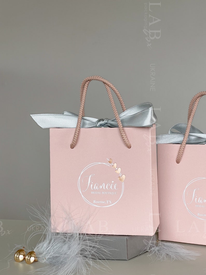 May include: Two pink gift bags with white rope handles and silver satin bows. The bag on the left has a logo that says "Fiancee Bridal Boutique, Boerne, TX".