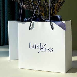 May include: Two white shopping bags with navy blue rope handles and navy blue ribbon. The bags have the word "Lush/ness" printed in navy blue. The bags are sitting on a white surface.