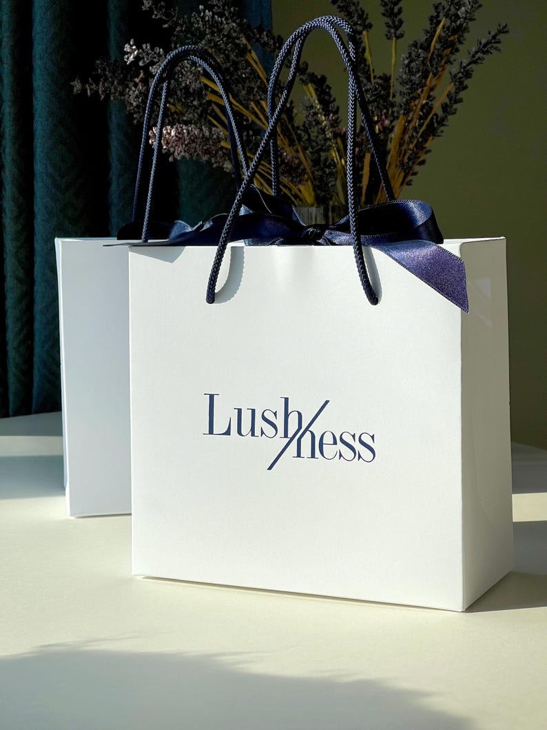 May include: Two white shopping bags with navy blue rope handles and navy blue ribbon. The bags have the word "Lush/ness" printed in navy blue. The bags are sitting on a white surface.