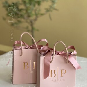 May include: Two pink gift bags with gold lettering. The bags have a gold logo that says "BP" and "BOLD PIECES" with the text "UKRAINE" below. The bags have pink satin ribbon handles.