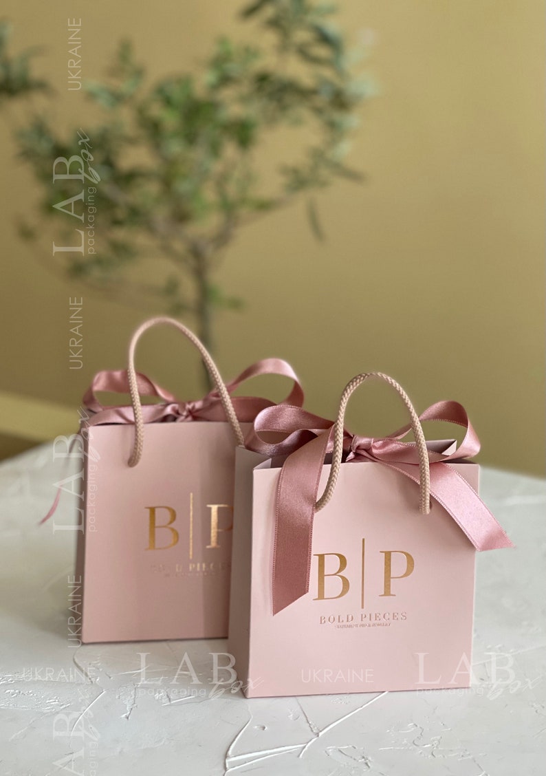 May include: Two pink gift bags with gold lettering. The bags have a gold logo that says "BP" and "BOLD PIECES" with the text "UKRAINE" below. The bags have pink satin ribbon handles.