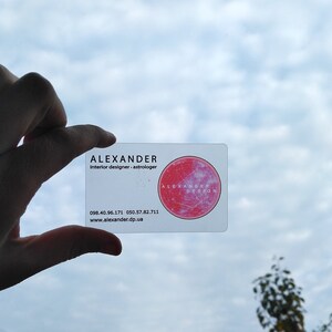 Op de afbeelding: Een witte visitekaartje met de tekst "ALEXANDER Interior designer.astrologer" en een rode cirkel met de tekst "ALEXANDER DESIGN" in wit. De telefoonnummers 098.40.96.171 en 050.57.82.711 en de website www.alexander.dp.ua staan ook op de kaart gedrukt.