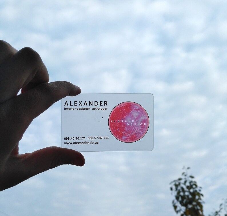 Op de afbeelding: Een witte visitekaartje met de tekst "ALEXANDER Interior designer.astrologer" en een rode cirkel met de tekst "ALEXANDER DESIGN" in wit. De telefoonnummers 098.40.96.171 en 050.57.82.711 en de website www.alexander.dp.ua staan ook op de kaart gedrukt.