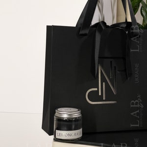 Puede incluir: Una bolsa de regalo negra con una cinta de sat&eacute;n negra y un logotipo plateado. La bolsa est&aacute; sobre una superficie blanca junto a un peque&ntilde;o frasco de vidrio con tapa plateada. El frasco contiene una sustancia negra y tiene una etiqueta blanca que dice "LEMONGRASS".