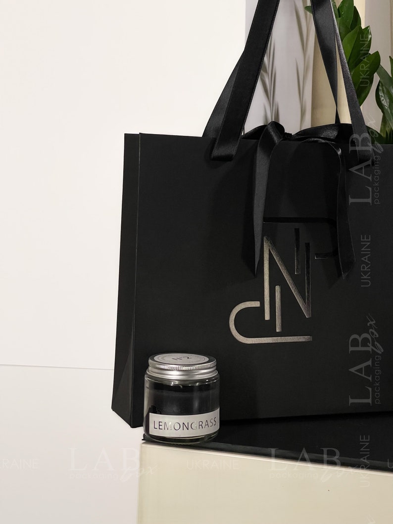 Puede incluir: Una bolsa de regalo negra con una cinta de sat&eacute;n negra y un logotipo plateado. La bolsa est&aacute; sobre una superficie blanca junto a un peque&ntilde;o frasco de vidrio con tapa plateada. El frasco contiene una sustancia negra y tiene una etiqueta blanca que dice "LEMONGRASS".