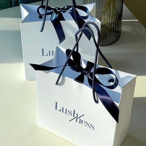 May include: Two white gift bags with navy blue ribbon bows and handles. The bags have the word "Lushness" printed on them in navy blue. The bags are sitting on a white surface.