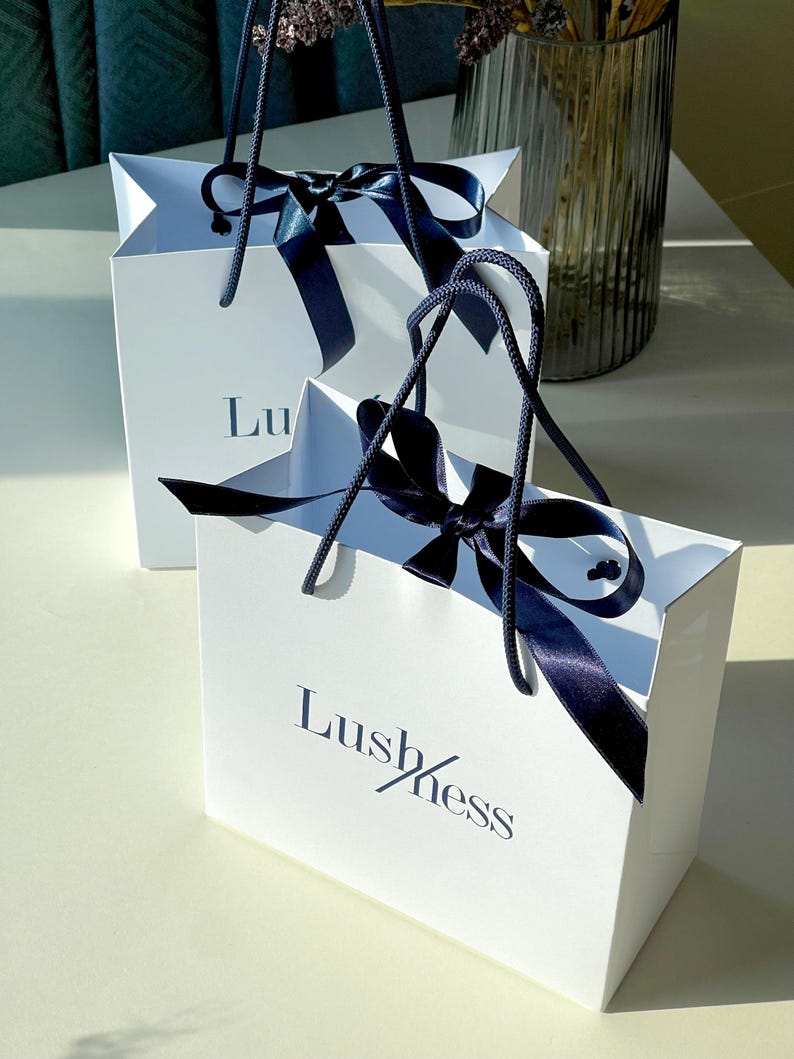 May include: Two white gift bags with navy blue ribbon bows and handles. The bags have the word "Lushness" printed on them in navy blue. The bags are sitting on a white surface.