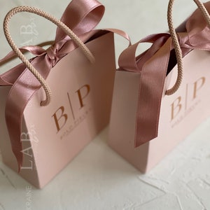 May include: Two pink gift bags with rope handles and a pink satin ribbon bow. The bags have the text "B|P" printed on the front.