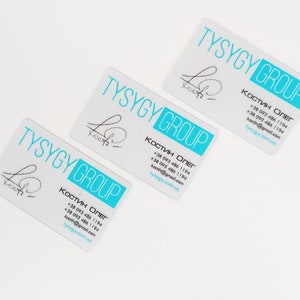Op de afbeelding: Drie witte visitekaartjes met blauwe tekst en een blauw logo. De tekst op de kaartjes luidt "TYSYGY GROUP" en bevat contactgegevens van Oleg Kostin. Het logo is een gestileerde handtekening.
