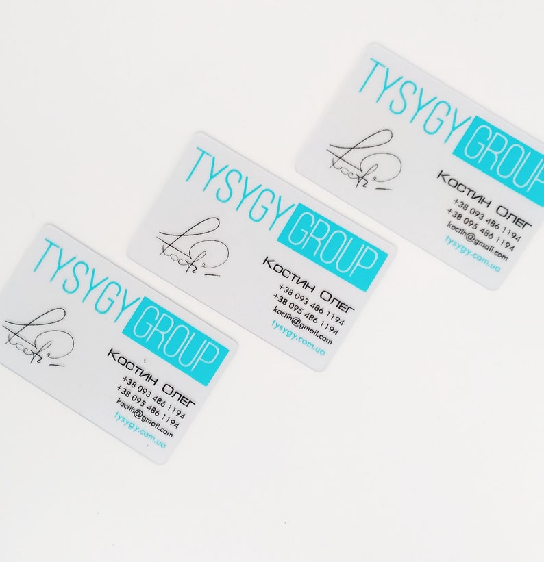 Op de afbeelding: Drie witte visitekaartjes met blauwe tekst en een blauw logo. De tekst op de kaartjes luidt "TYSYGY GROUP" en bevat contactgegevens van Oleg Kostin. Het logo is een gestileerde handtekening.