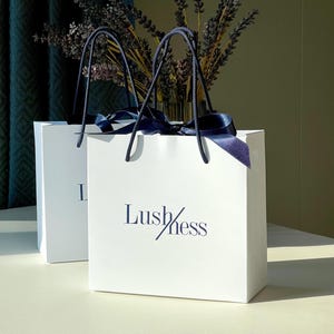Pode incluir: Duas sacolas de presente brancas com alças e fitas azul marinho. As sacolas têm a palavra "Lushness" impressa em azul marinho. Um vaso de lavanda seca é visível ao fundo.