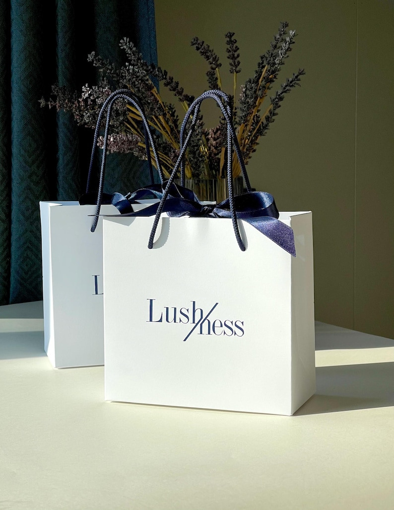 May include: Two white gift bags with navy blue handles and ribbon. The bags have the word "Lushness" printed in navy blue. A vase of dried lavender is visible in the background.