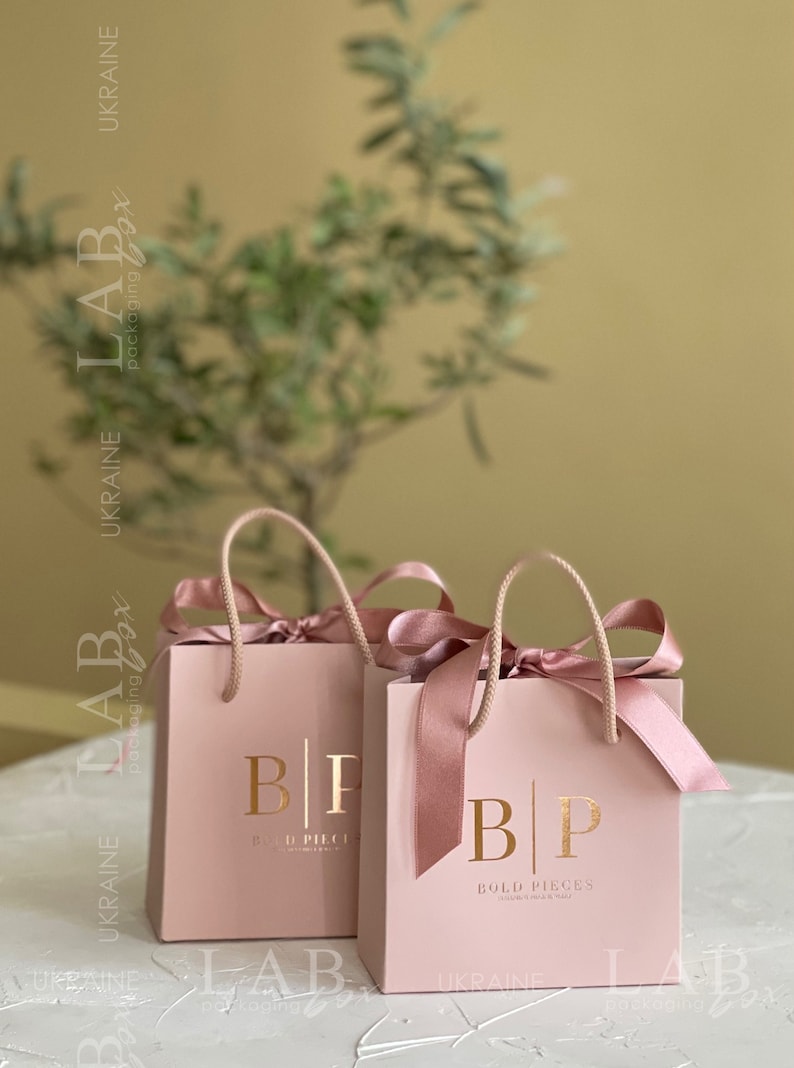 May include: Two pink gift bags with gold lettering that reads "Bold Pieces" and a matching pink ribbon. The bags are on a white surface.