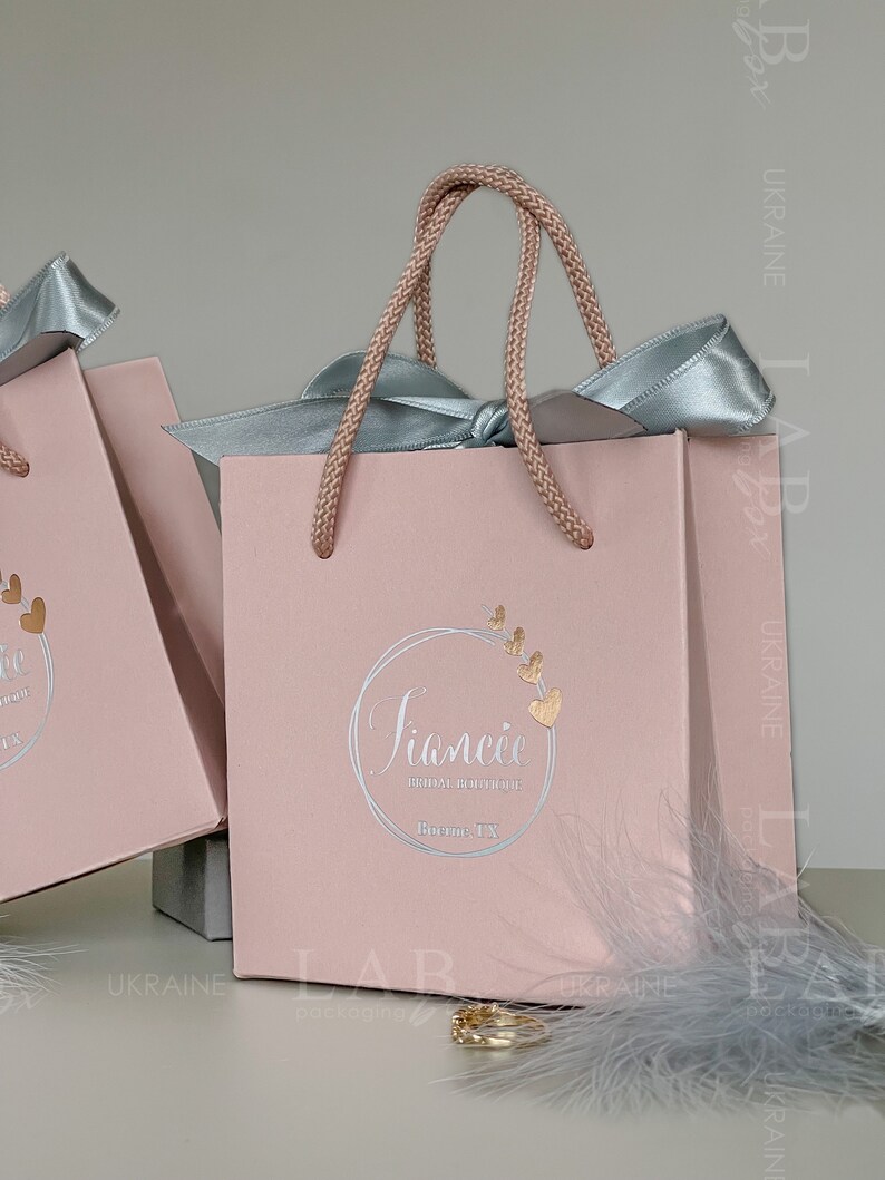 May include: A pink gift bag with a light blue ribbon and a gold logo that says "Siancie Bridal Boutique Bourne, TX".