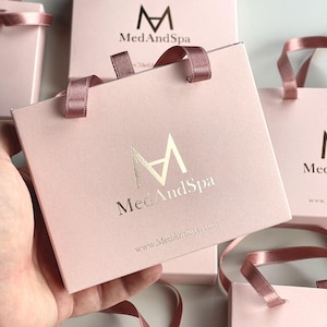 May include: Pink gift bags with matching ribbon handles. The bags feature the logo "M" above the words "MedAndSpa" in gold lettering. The website address www.MedAndSpa.com is printed below the logo. Several bags are visible.