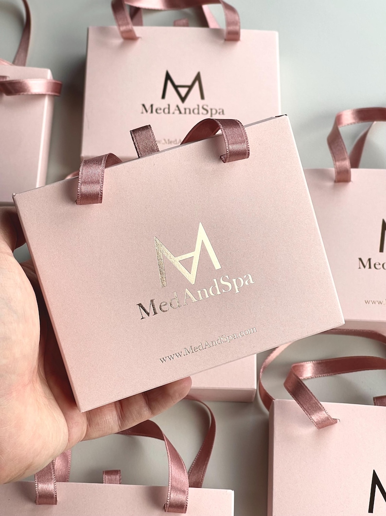 May include: Pink gift bags with matching ribbon handles. The bags feature the logo "M" above the words "MedAndSpa" in gold lettering. The website address www.MedAndSpa.com is printed below the logo. Several bags are visible.