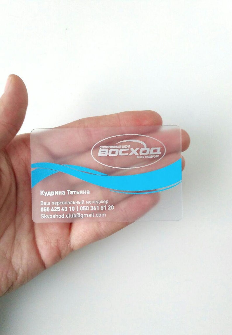 Op de afbeelding: Een transparante plastic visitekaartje met witte tekst en een blauw logo. De tekst luidt "Спортивный клуб Восход быть лидером?" en "Кудрина Татьяна Ваш персональный менеджер 050 425 43 10 | 050 361 51 20 Skvoshod.club@gmail.com".