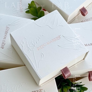 Pode incluir: Caixas de presente brancas com um design floral em relevo e o texto "DEPO MARKET" em letras douradas rosadas. As caixas têm um puxador de fita rosa.
