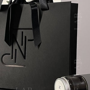 Puede incluir: Una bolsa de regalo negra con una cinta de sat&eacute;n negra y un dise&ntilde;o recortado de la letra "N". La bolsa est&aacute; sobre una superficie negra junto a un peque&ntilde;o frasco de vidrio con tapa negra. El frasco tiene una etiqueta blanca con el texto "N&deg;2" y "ALMOND & VANILLA" impreso en ella.
