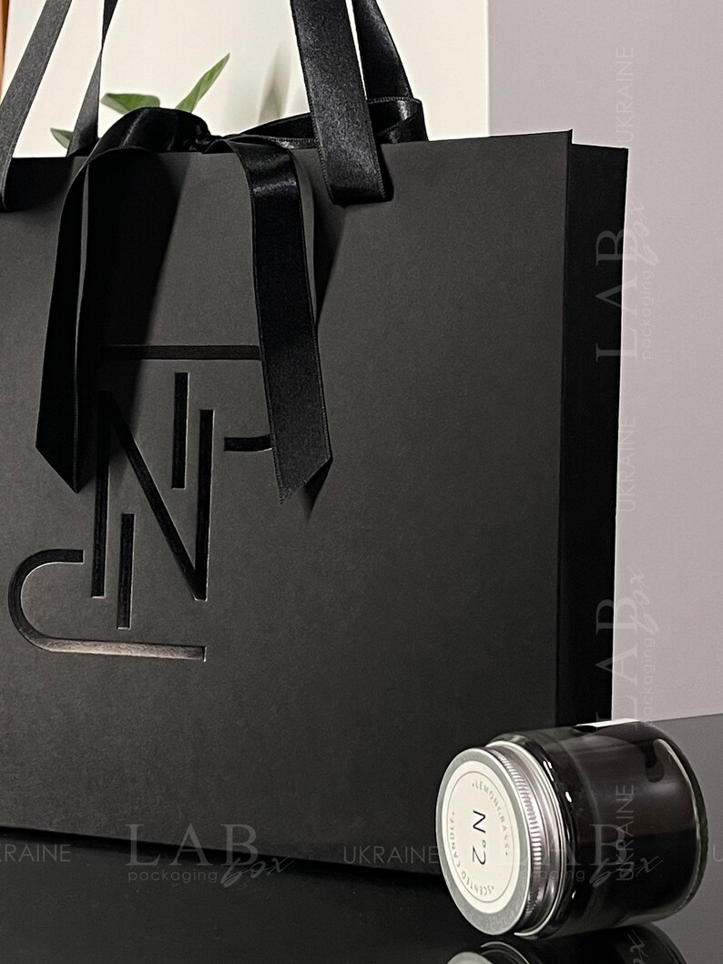 Puede incluir: Una bolsa de regalo negra con una cinta de sat&eacute;n negra y un dise&ntilde;o recortado de la letra "N". La bolsa est&aacute; sobre una superficie negra junto a un peque&ntilde;o frasco de vidrio con tapa negra. El frasco tiene una etiqueta blanca con el texto "N&deg;2" y "ALMOND & VANILLA" impreso en ella.
