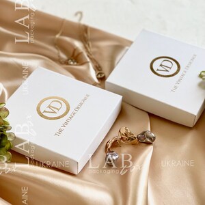 May include: Two white square boxes with gold lettering. The boxes have the text "The Vintage Designer" and the initials "VD" in a circle. The boxes are on a light brown satin surface.