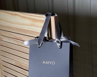 May include: A gray gift bag with a gray satin ribbon and the word "AMYO" printed in gold on the front.