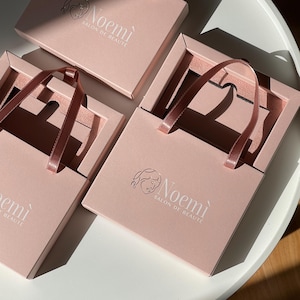 Puede incluir: Bolsas de regalo rosas con asas de cinta de raso. Las bolsas son rectangulares con un recorte en la parte superior. El texto "Noemi Salon de Beaute" está impreso en la parte delantera de las bolsas. Están dispuestas sobre una superficie blanca.