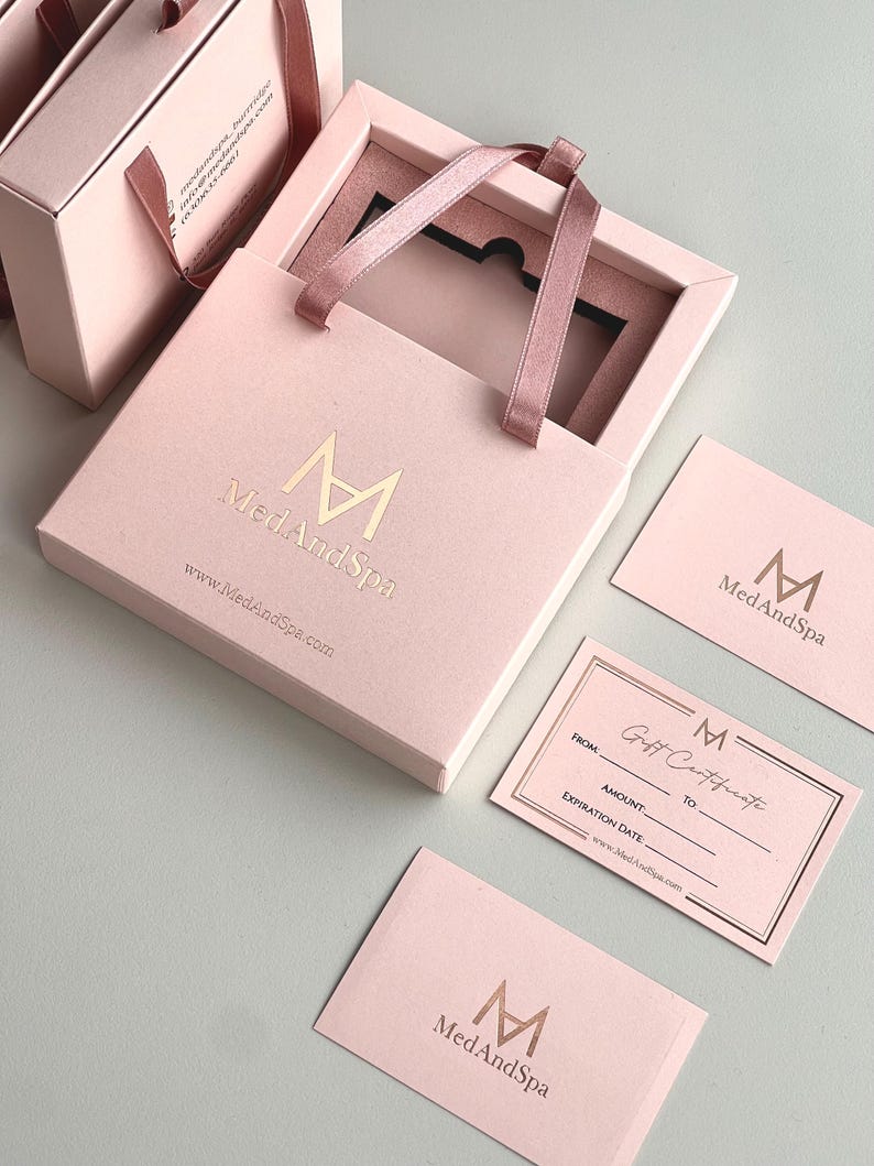 May include: Pink gift boxes and cards with gold foil lettering. The boxes have ribbon handles and the text "MedAndSpa" and website address. One card is a gift certificate with fields for recipient and amount.