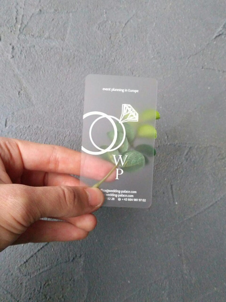 Op de afbeelding: Een mat plastic visitekaartje met een wit ring- en diamantontwerp. De tekst op de kaart luidt "event planning in Europe", "W P", "info@wedding-palace.com", "wedding-palace.com", "82 28" en "+43 664 981 97 02".