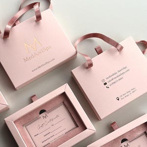 May include: Pink gift boxes with ribbon handles and the "MedAndSpa" logo in gold. Some boxes are open, revealing gift certificates. The gift certificates have space for "From," "Amount," and "Expiration Date."