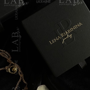 May include: A black jewelry box with a gold embossed logo that reads "LENA RIABININA Jewelry". The box is on a black velvet surface.