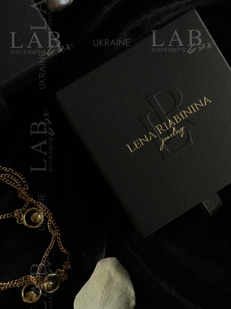 May include: A black jewelry box with a gold embossed logo that reads "LENA RIABININA Jewelry". The box is on a black velvet surface.