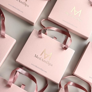 May include: Pink gift bags with matching ribbon handles. The bags have a gold logo that reads "Med And Spa" and the website address. The bags are arranged on a light gray surface.
