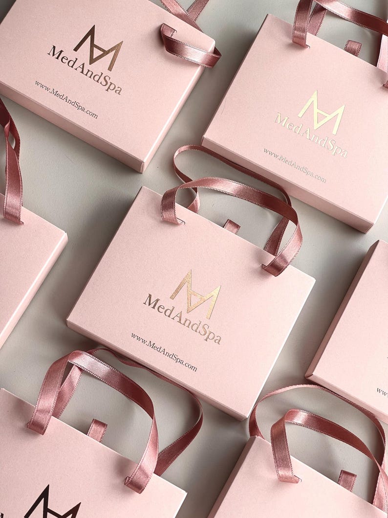May include: Pink gift bags with matching ribbon handles. The bags have a gold logo that reads "Med And Spa" and the website address. The bags are arranged on a light gray surface.