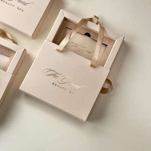 May include: Pale pink gift boxes with gold lettering reading "The Grand Beauty Spa". Each box has a matching ribbon handle and a gift card inside. The boxes are arranged on a white surface.
