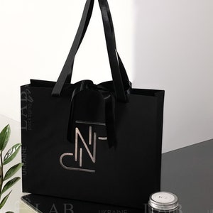 Puede incluir: Una bolsa de regalo negra con asas de sat&eacute;n negro y un lazo de sat&eacute;n negro. La bolsa tiene un logotipo plateado que dice "N-". Un peque&ntilde;o frasco de vidrio con tapa plateada y una etiqueta blanca que dice "N2" y "MONGRASS" est&aacute; sentado sobre una superficie negra.