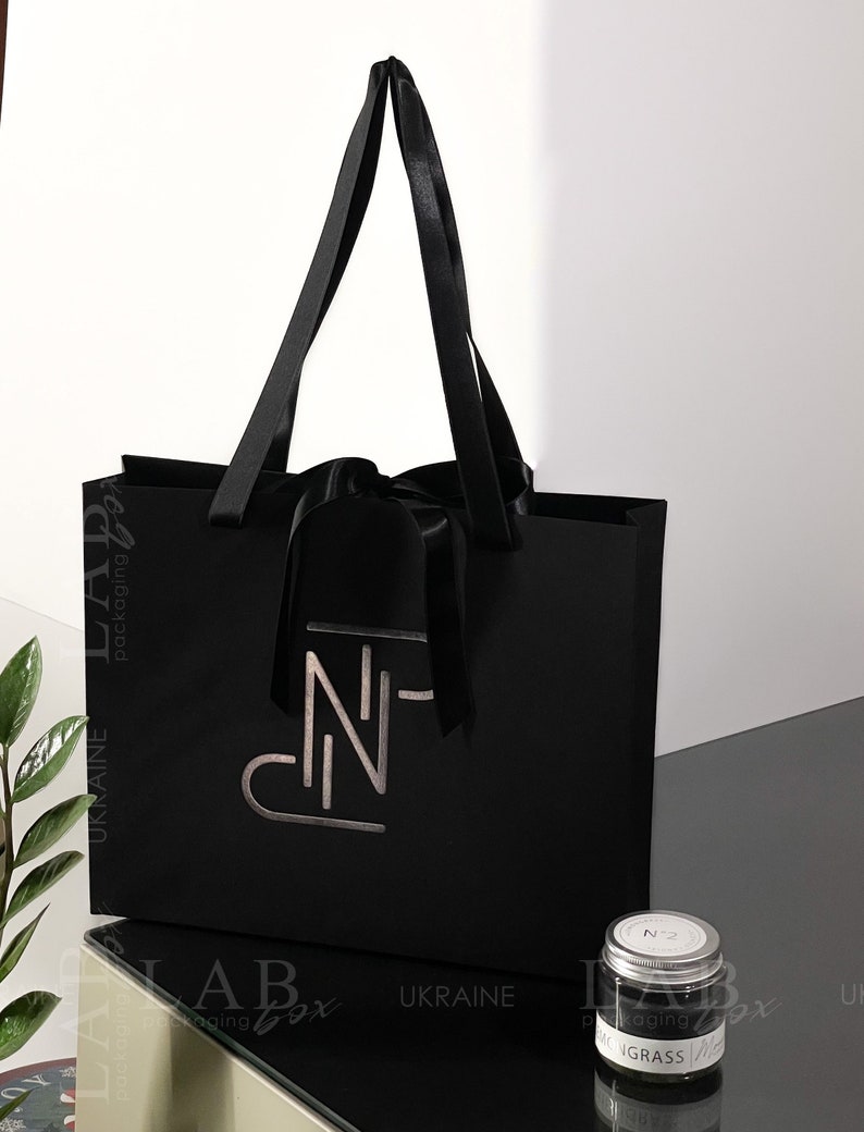 Puede incluir: Una bolsa de regalo negra con asas de sat&eacute;n negro y un lazo de sat&eacute;n negro. La bolsa tiene un logotipo plateado que dice "N-". Un peque&ntilde;o frasco de vidrio con tapa plateada y una etiqueta blanca que dice "N2" y "MONGRASS" est&aacute; sentado sobre una superficie negra.