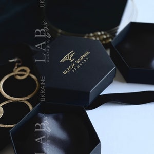 Puede incluir: Dos cajas hexagonales negras con un logotipo dorado que dice "BLACK SAVANNA JEWELRY". Las cajas están sobre una superficie negra con una cinta negra.