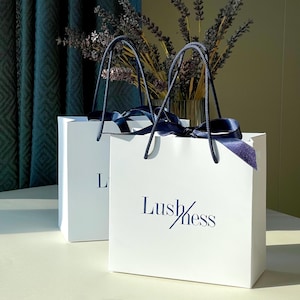 May include: Two white gift bags with the word "Lushness" printed in navy blue. The bags have navy blue handles and are tied with navy blue ribbons. Dried lavender is in a clear vase in the background.